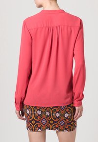 Korálová blůza s dlouhými rukávy a skrytými sklady na zádech, v kombinaci s vzorovanou sukní s geometrickými motivy v červené, žluté a bílé na tmavém pozadí.