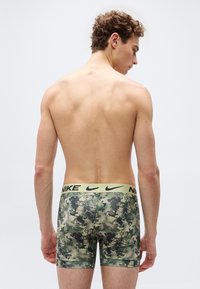 Άνδρας μοντέλο εμφανίζεται από πίσω φορώντας πράσινα καμουφλάζ μποξεράκια Nike με κίτρινη ζώνη μέσης, σε απλό φωτεινό φόντο.