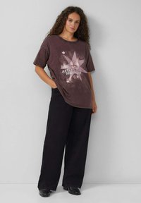 Übergroßes, dunkelviolettes T-Shirt mit grafischen Sternen und dem Text "NACH Mitternacht", kombiniert mit schwarzen, weit geschnittenen Hosen und schwarzen Schuhen.