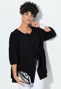 Black peplum top with three-quarter puff sleeves, textured fabric, tied front closure, paired with a silver lip-shaped clutch.