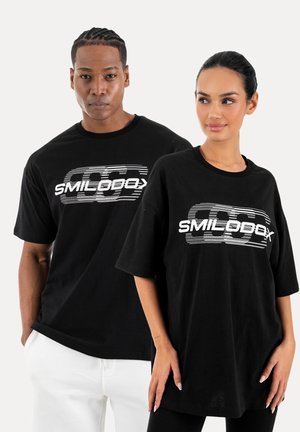 Mand og kvinde står side om side, begge iført sorte oversized T-shirts med hvide "SMILODOX" logoer foran.