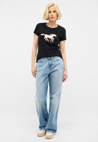 Camiseta negra con un gráfico de caballo y texto "Mustang", combinada con pantalones de mezclilla anchos azul claro y zapatillas beige.
