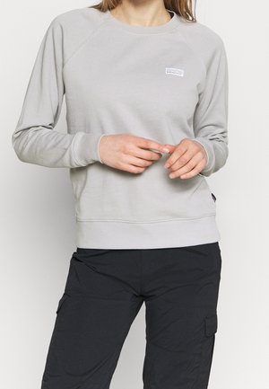 Kobieta w jasnoszarej bluzie Patagonia i czarnych spodniach typu cargo, dłonie złączone w pobliżu talii, jednolite tło