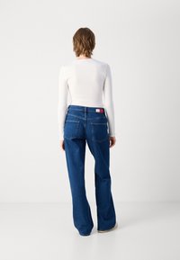 Blue denim jeans with a straight leg, two back pockets, and a branded waistband tag. Paired with a white long-sleeve top.