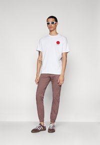Witte T-shirt met een rode cirkelafbeelding, gecombineerd met lichtbruine tapered broek en bruine sneakers met witte strepen. Witte zonnebril.