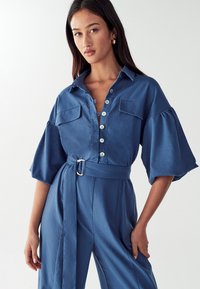 Kvinna klädd i en blå jumpsuit med bälte, vida ärmar och knappar fram, stående mot en enfärgad ljus bakgrund.