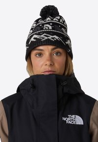 Beanie lavorato a maglia in bianco e nero con pom-pom e motivi geometrici. Abbinato a un giubbotto nero e talpa con un logo sul petto.