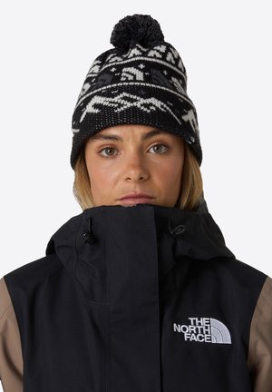 Czarny i biały dzianinowy beanie z pomponem i wzorami geometrycznymi. Połączony z czarną kurtką w odcieniu taupe z logo na piersi.
