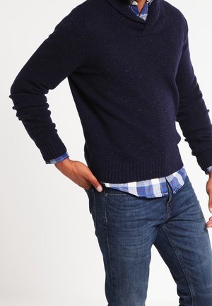 Man met een donkerblauw gebreide trui over een blauw-wit geruit overhemd en een blauwe spijkerbroek, hand in de zak, tegen een witte achtergrond.