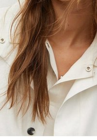 Witte, knopen-up jas met hoge kraag, voorzien van zilveren drukkers en een gladde, gestructureerde stof; het bruine haar van het model is gedeeltelijk zichtbaar.