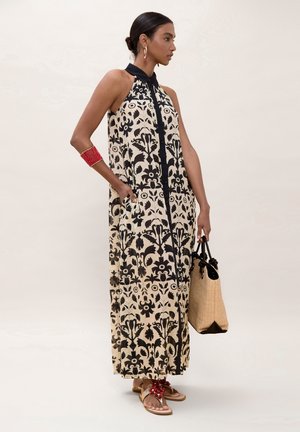 Mujer con vestido halter floral en negro y beige, con pulsera roja y pendientes, sosteniendo un bolso tote tejido beige.