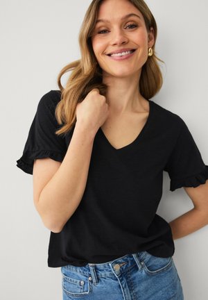 Smiling woman with shoulder-length blonde hair wearing a black ruffled sleeve V-neck shirt and blue jeans, posing with one hand near her neck.