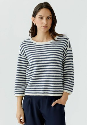 Femme aux cheveux foncés portant un pull à rayures horizontales blanches et bleu marine avec des manches trois-quarts et un pantalon bleu marine, une main dans la poche.