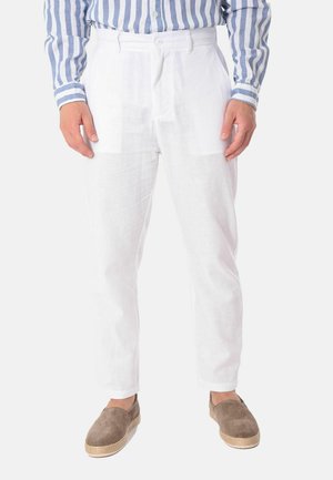 Uomo che indossa pantaloni di lino bianchi, una camicia a righe blu e bianca e scarpe marroni senza lacci, in piedi davanti a uno sfondo bianco.