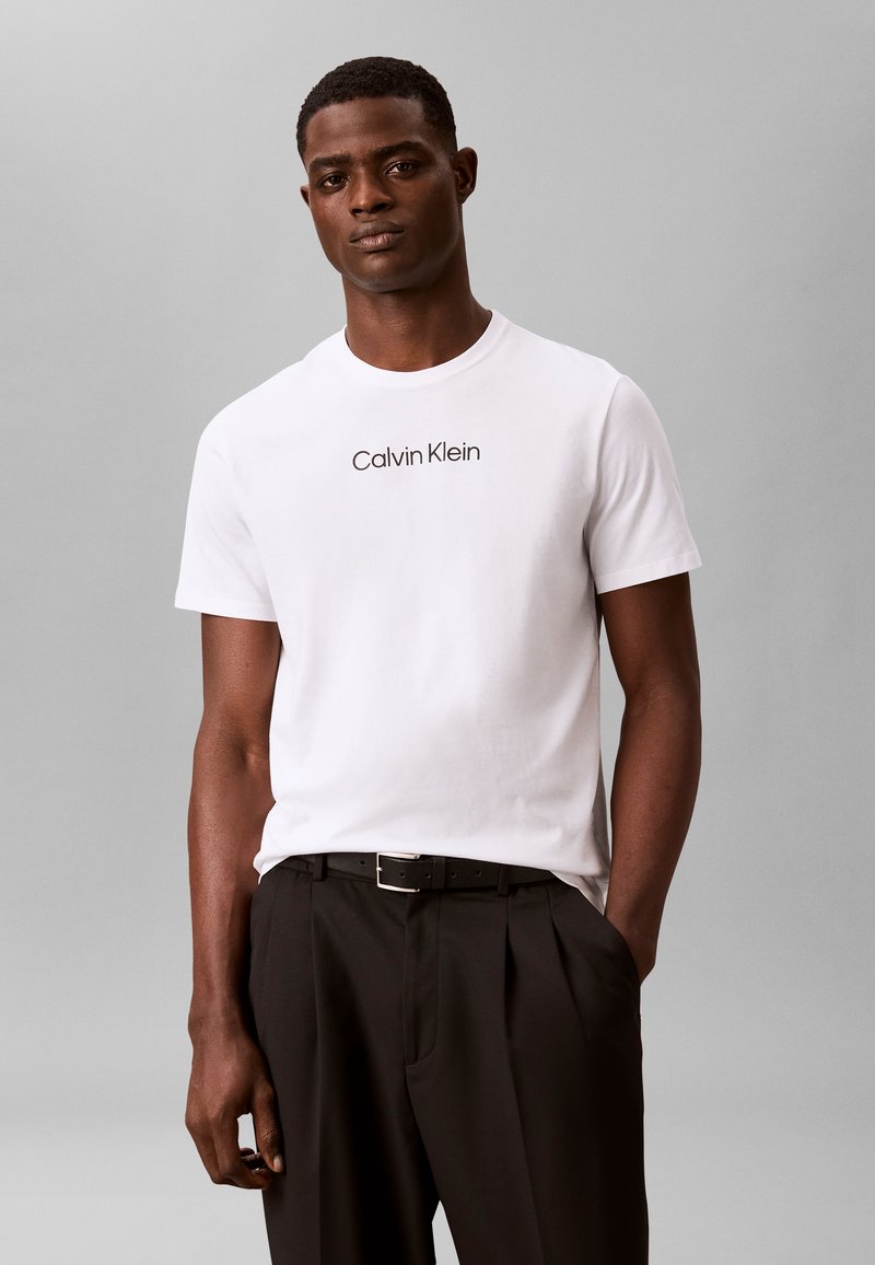 Άνδρας που φοράει λευκό t-shirt Calvin Klein και μαύρο παντελόνι με τα χέρια στις τσέπες, στέκεται μπροστά σε ένα απλό γκρίζο φόντο.