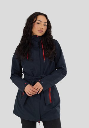 Donna che indossa un impermeabile blu navy con dettagli rossi sulla cerniera e cintura annodata in vita, in piedi davanti a uno sfondo grigio chiaro uniforme.