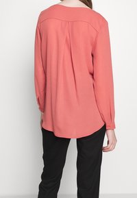 Femme portant un chemisier corail à manches longues avec un détail plis au dos, associé à un pantalon noir, vue de dos sur un fond blanc uni.