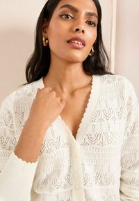 White knitted cardigan featuring a scalloped neckline and patterned texture with zigzag and leaf motifs, complemented by button closure.