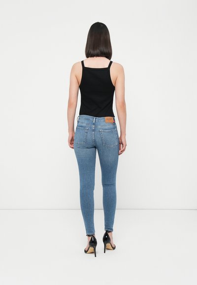 Jeans skinny bleu avec un délavé moyen, débardeur noir et chaussures à talons noirs ; la vue arrière montre deux poches arrière et un détail d'étiquette.