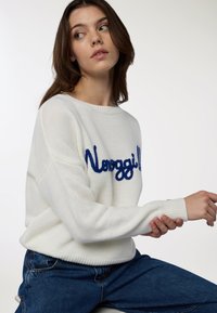 Maglione bianco in maglia con scollo rotondo e maniche lunghe, caratterizzato da testo ricamato in blu sul davanti, abbinato a jeans di denim.