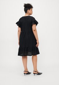 Black eyelet dress with ruffled sleeves, composed of textured fabric, featuring a tiered hem and a relaxed fit. Worn with black heeled shoes.