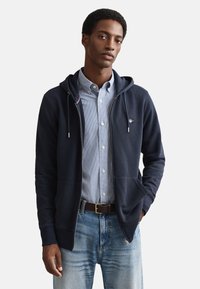 Felpa con zip blu navy in misto cotone, dotata di cappuccio con coulisse, due tasche frontali e un piccolo logo sul petto. Abbinata a una maglietta a righe.