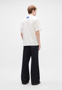 Λευκό βαμβακερό μπλουζάκι με μπλε ετικέτα στο πίσω μέρος, συνδυασμένο με σκούρους φαρδιούς τζιν παντελόνες. Το σύνολο διαθέτει άνετη εφαρμογή και μινιμαλιστικό σχέδιο.