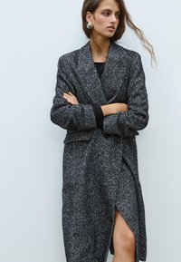Cappotto lungo grigio scuro con motivo a spina di pesce, vestibilità over, ampi rever e chiusura con un solo bottone. Dotato di uno spacco laterale e tessuto strutturato.