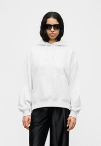 Γκρι φαρδιά φούτερ με τσέπη μπροστά, κορδόνια και ριμπ τελείωμα, συνδυασμένο με γυαλιστερό μαύρο παντελόνι. Το μοντέλο φοράει μαύρα γυαλιά ηλίου.