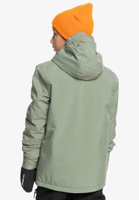 Zelená voděodolná bunda s kapucí, texturovaný materiál a černé rukavice. Pozadí je bílé, což zdůrazňuje detaily oblečení.