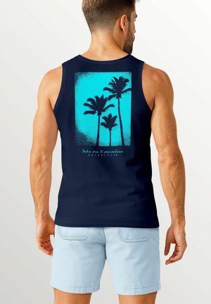 Ein Mann in einem marineblauen Tanktop mit einem Palmenmotiv auf dem Rücken steht vor einem hellen Hintergrund und trägt hellblaue Shorts.