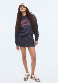 Marinblå sweatshirt med röd text "BROOKLYN NEW YORK", kombinerad med en mörk denim mini kjol och mönstrade bruna sneakers.