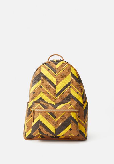 Sac à dos marron, jaune et noir avec un motif en chevrons, présentant une poche zippée à l'avant et une finition texturée. Accents de logo visibles.