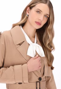 Beiger Trenchcoat mit breitem Kragen, weißer Fliege und geknöpften Manschetten. Auffällige Details sind ein taillierter Gürtel und dunkle Knöpfe.