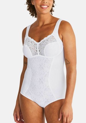Lachende vrouw die een wit kanten body draagt met brede bandjes, bloemmotieven en een tatoeage op de bovenarmen tegen een effen achtergrond.