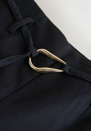 Detailní záběr tmavě modré látky s texturovaným povrchem, zdobené zlatým kovovým srdíčkem a předeným látkovým detailem.