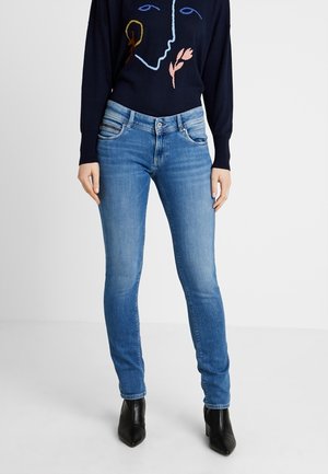 Osoba nosząca dopasowane niebieskie dżinsy, czarne botki do kostki oraz granatowy sweter z abstrakcyjną twarzą i kwiatowym wzorem.