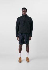 Schwarze Fleecejacke mit hohem Kragen, Reißverschluss vorne und Logodetail, kombiniert mit navy-blauen Cargoshorts und beigen Sneakers. Weiße Socken inklusive.