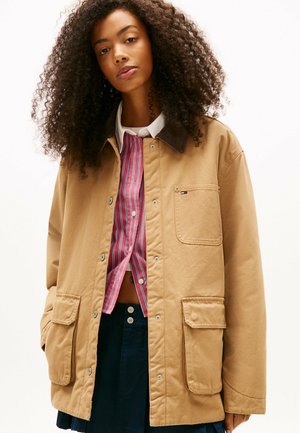 Chaqueta de algodón color beige con cuello de pana marrón, cierres a presión, dos bolsillos frontales, llevada sobre una camisa a rayas rojas y blancas, combinada con una falda azul marino.