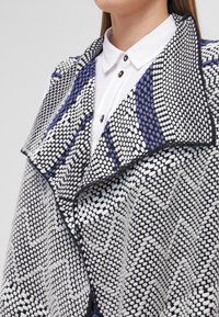 Cappotto a maglia con ampio collo in motivi nero, bianco e blu navy. Presenta un tessuto testurizzato e chiusura con bottone al collo.