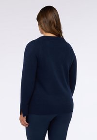 Una donna con capelli castani indossa un maglione a maniche lunghe blu scuro e pantaloni abbinati, in piedi con la schiena rivolta verso la fotocamera su uno sfondo bianco.