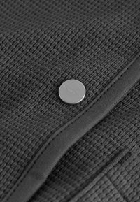 Dark gray textured fabric with a waffle pattern, featuring a silver button with the engraving "ALLSAINTS" and a smooth contrasting trim.