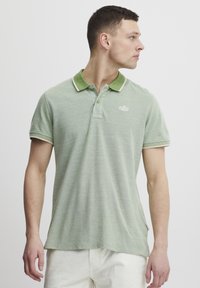 Grønn polo skjorte med teksturert stoff, kontrasterende grønn krage og hvit piping. Har en liten logo på brystet og korte ermer.
