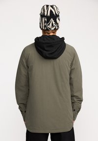 Veste en velours c�ôtelé vert olive avec une capuche noire, présentant un tissu texturé et des détails de boutons-pression aux poignets. Bonnet à motifs noir et blanc.