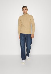 Suéter de cuello alto de punto beige con un diseño de cable texturizado, combinado con vaqueros sueltos de color azul oscuro y zapatillas blancas.