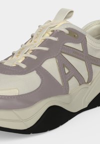 Κοντινό πλάνο ενός κρεμ και μάβ sneaker με παχιά σόλα και κορδόνια, με το λογότυπο "Armani Exchange" στην πλευρά και στο loop της γλώσσας.