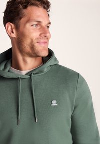 Man with short curly hair wearing green hooded sweatshirt with logo, looking slightly to the right against a plain background.