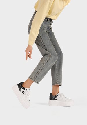 Sneakers bianchi con tallone in pelle nera, suola platform e design con lacci, indossati con jeans grigi a gamba corta e una maglietta gialla.