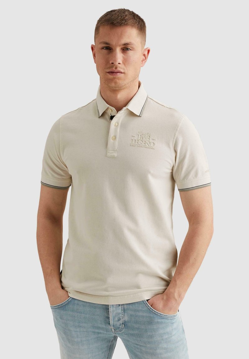 Jeune homme portant un polo beige avec un logo brodé et un jean bleu clair, debout les mains dans les poches devant un fond uni.