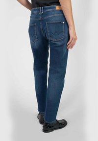 Persoon in donkerblauwe rechtzittende jeans en zwarte glanzende loafers, staand met één hand ontspannen naast hun lichaam.